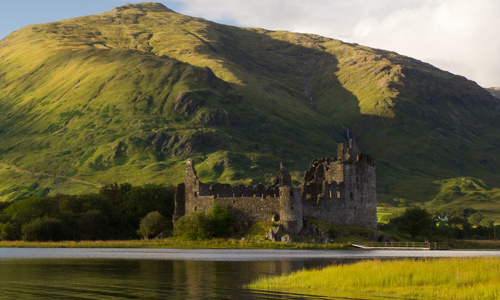 The very picturesque Kilchurn Castle viewed from Loch Awe, with sun-touched hills just behind it.