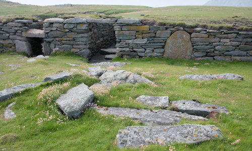 A stone wall that used to be part of a house, with stone slabs on the ground in front of it.
