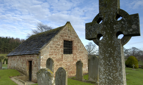 A celtic cross in front of a small stone house