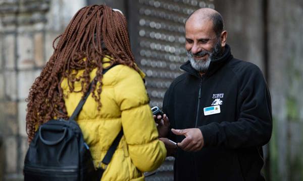 A man with a beard and a name badge smiles at a person in a yellow coat as he scans a ticket.