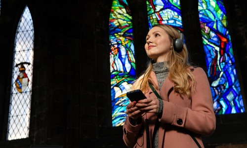 A person standing in front of a stained glass window smiling, holding an audio guide