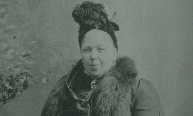 A woman in Victorian clothing and a feathered hat looks at the camera, slightly laughing