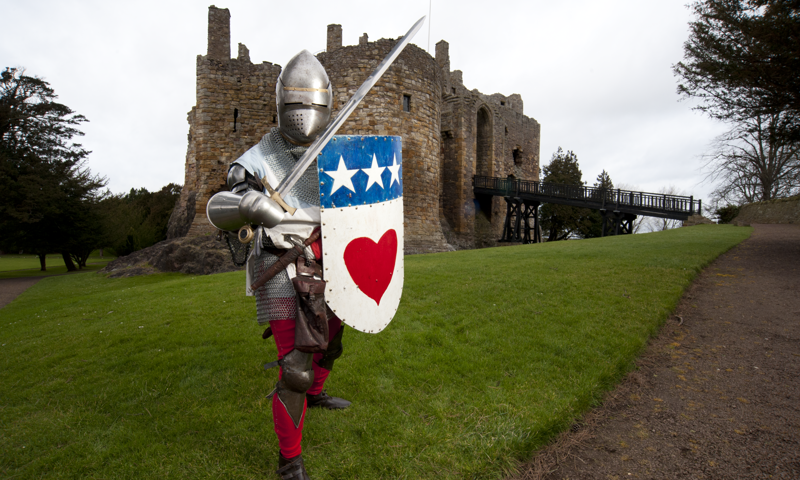 Knight in armour holding a sword and shield in a defensive position is standing on the grass in front of Dirleton Castle.