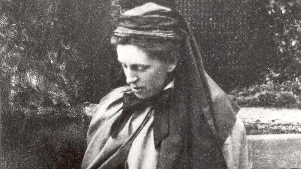 Here is concise, neutral alt text:  > Black-and-white photograph of a person wearing layered clothing and a headscarf, shown in profile outdoors, with foliage and a path in the background.
