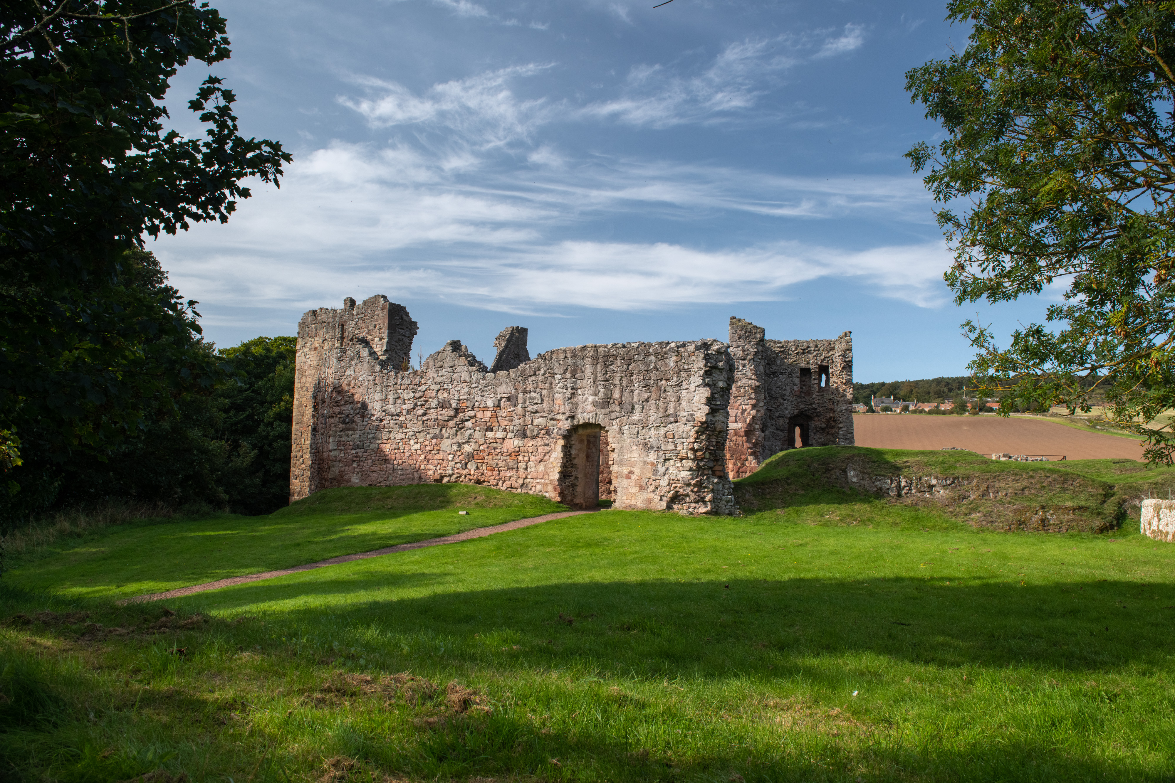 Ground-level view of Hailes Castle ruins with broken stone walls and arched openings, surrounded by green grass and framed by trees.