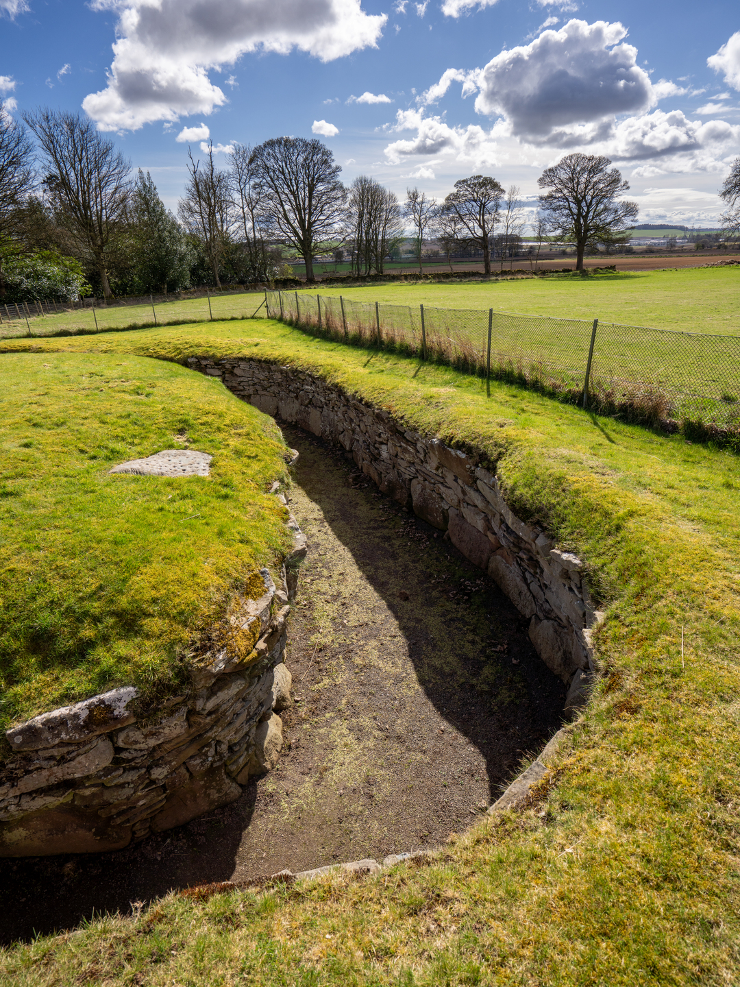 Part of a long, thin passage, with stones either side. There is grass on the ground above it and a fence surrounding it.