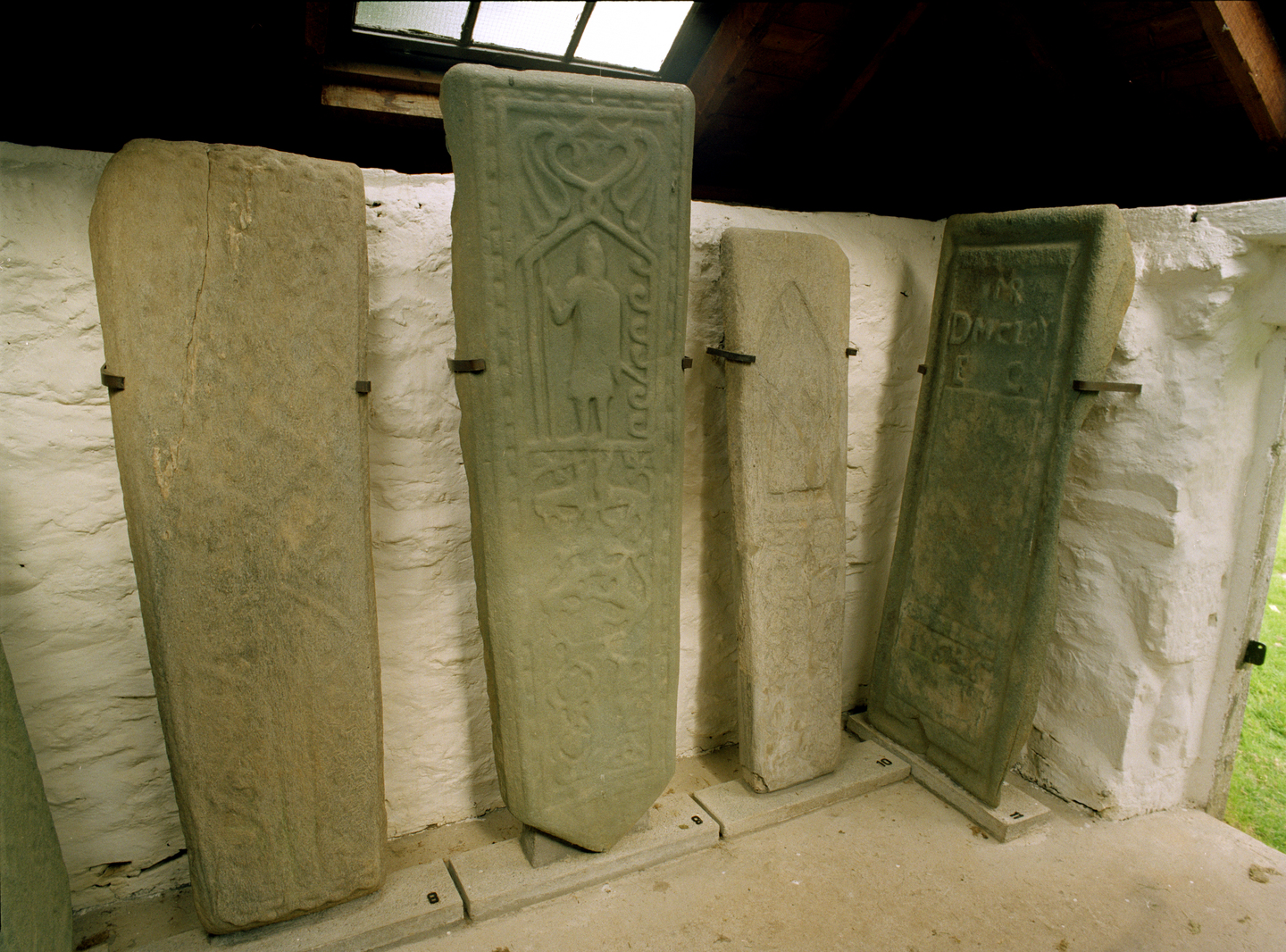 Four standing stone sculptures lined up. They are of various sizes, all vaguely rectangular. There are faded engravings on them, one has letters, another has the figure of a person. They are a grey colour, some with green staining.