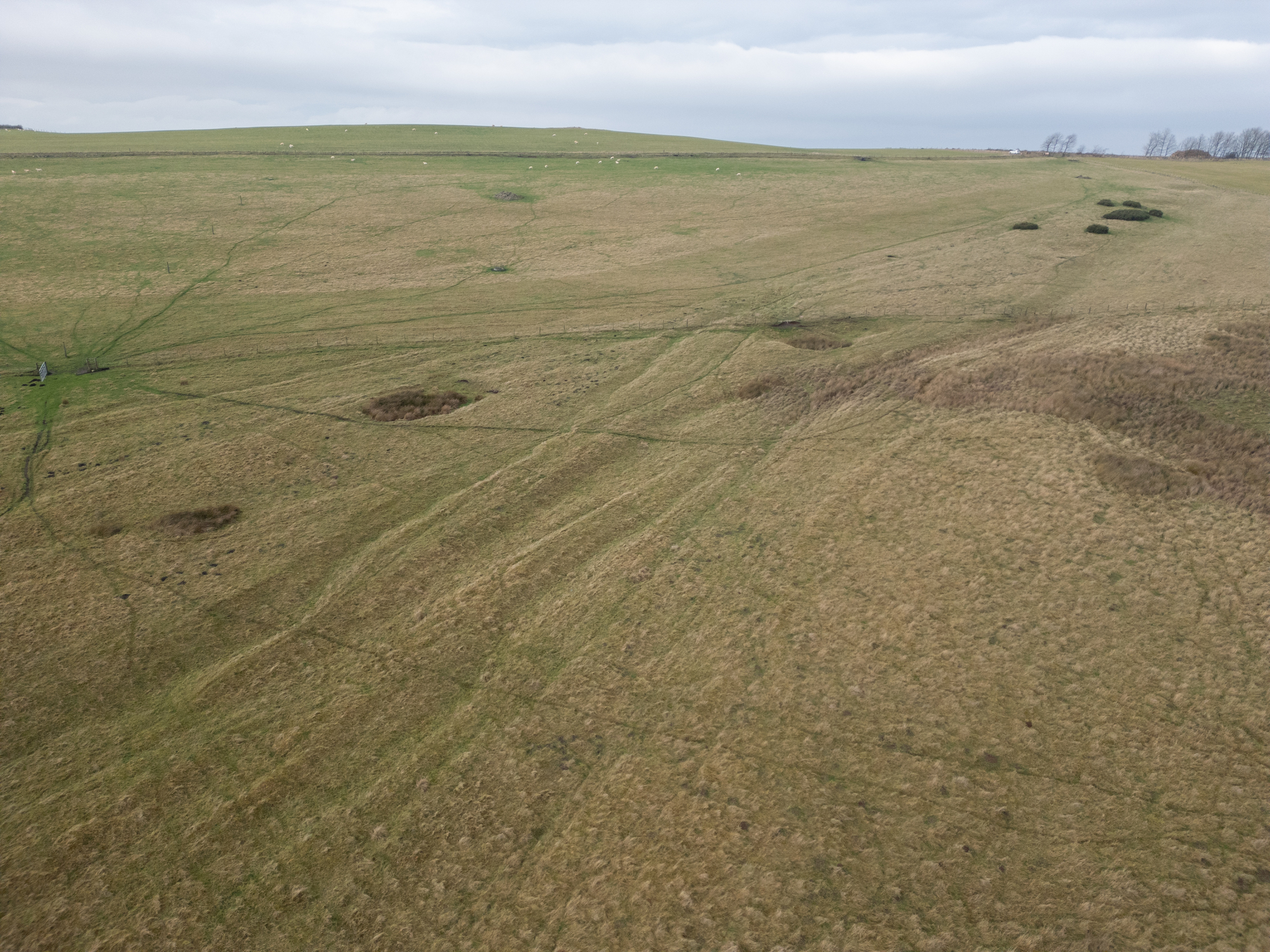 Aerial view of Dere Street Roman Road as faint linear earthworks running across an open grassy moorland. The landscape shows subtle ridges and depressions marking the ancient route, with scattered patches of darker vegetation and a cloudy sky above.