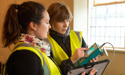 A photograph of two women with yellow hi vis jackets on, both are holding and looking at equipment. They are standing in a bright room together.