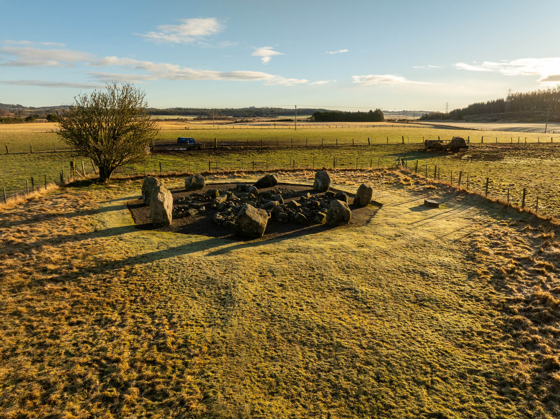 Wide view of the stone circle at dawn, with long shadows cast by the standing stones across frosty grass and open farmland stretching into the distance.