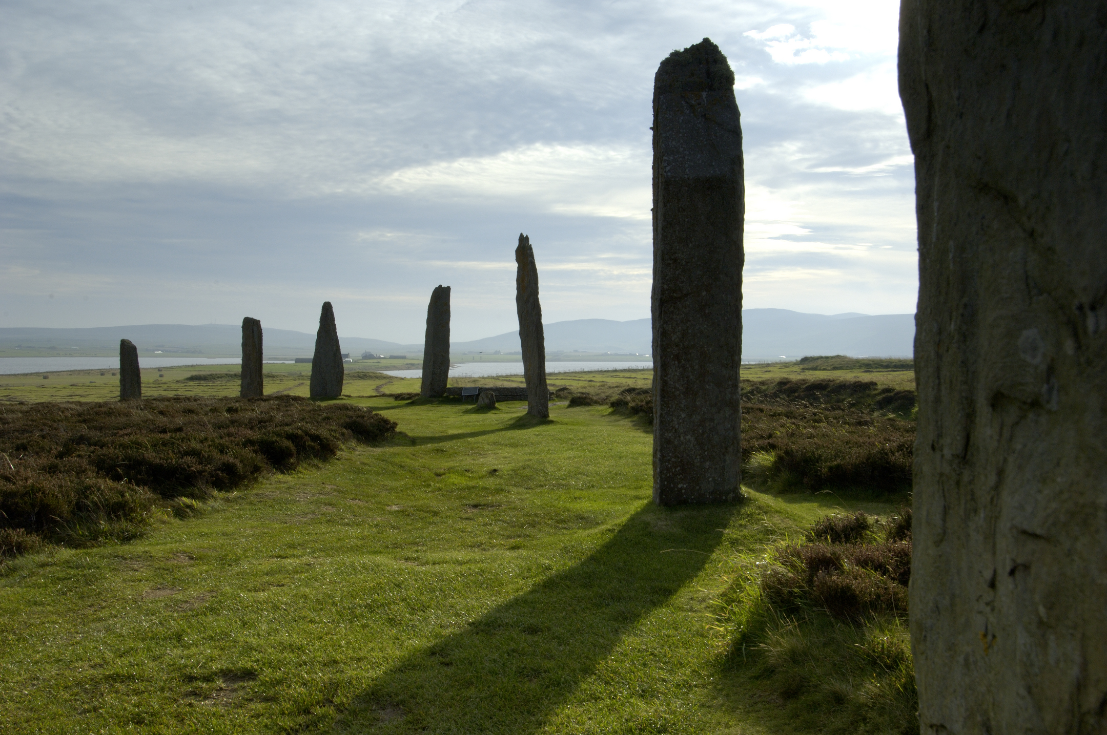 A large standing stone casts a shadow over a well-trodden grassy path. 