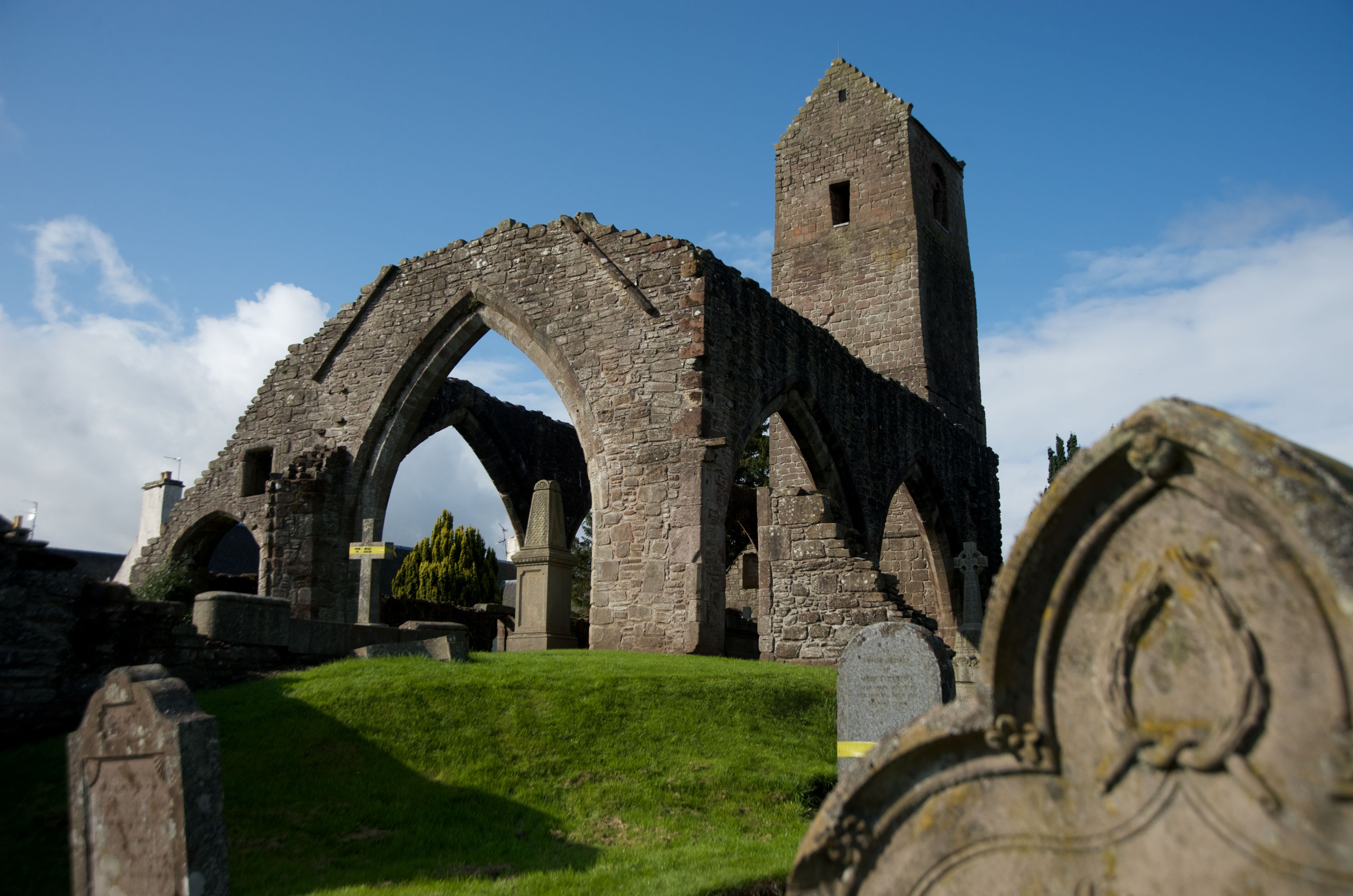 A wide view of the ruined Muthill Old Church, with its tall stone tower and pointed arches rising above a grassy churchyard dotted with gravestones.