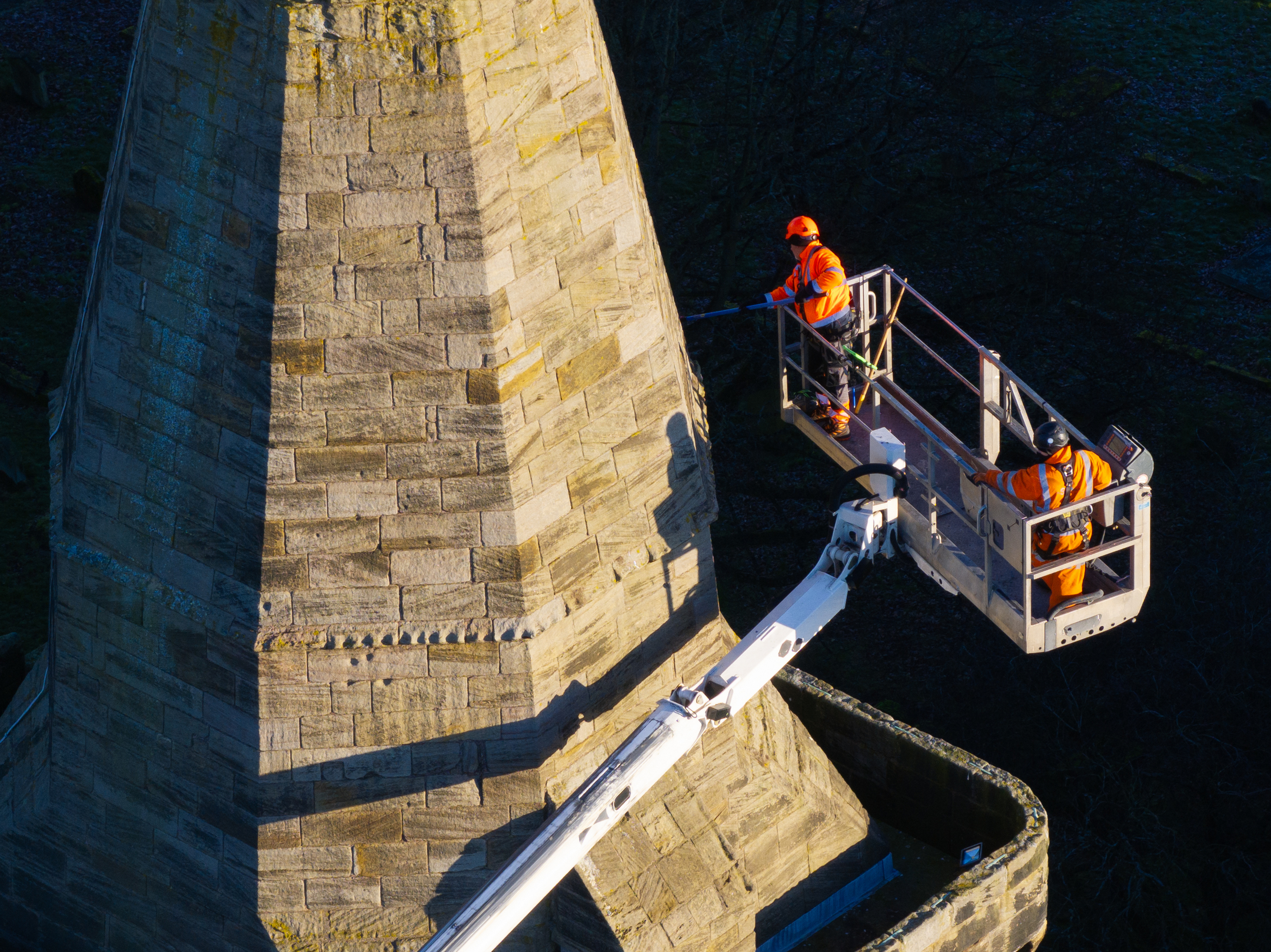 Two workers in safety gear operate a raised mechanical platform beside the steep stone tower of an abbey.