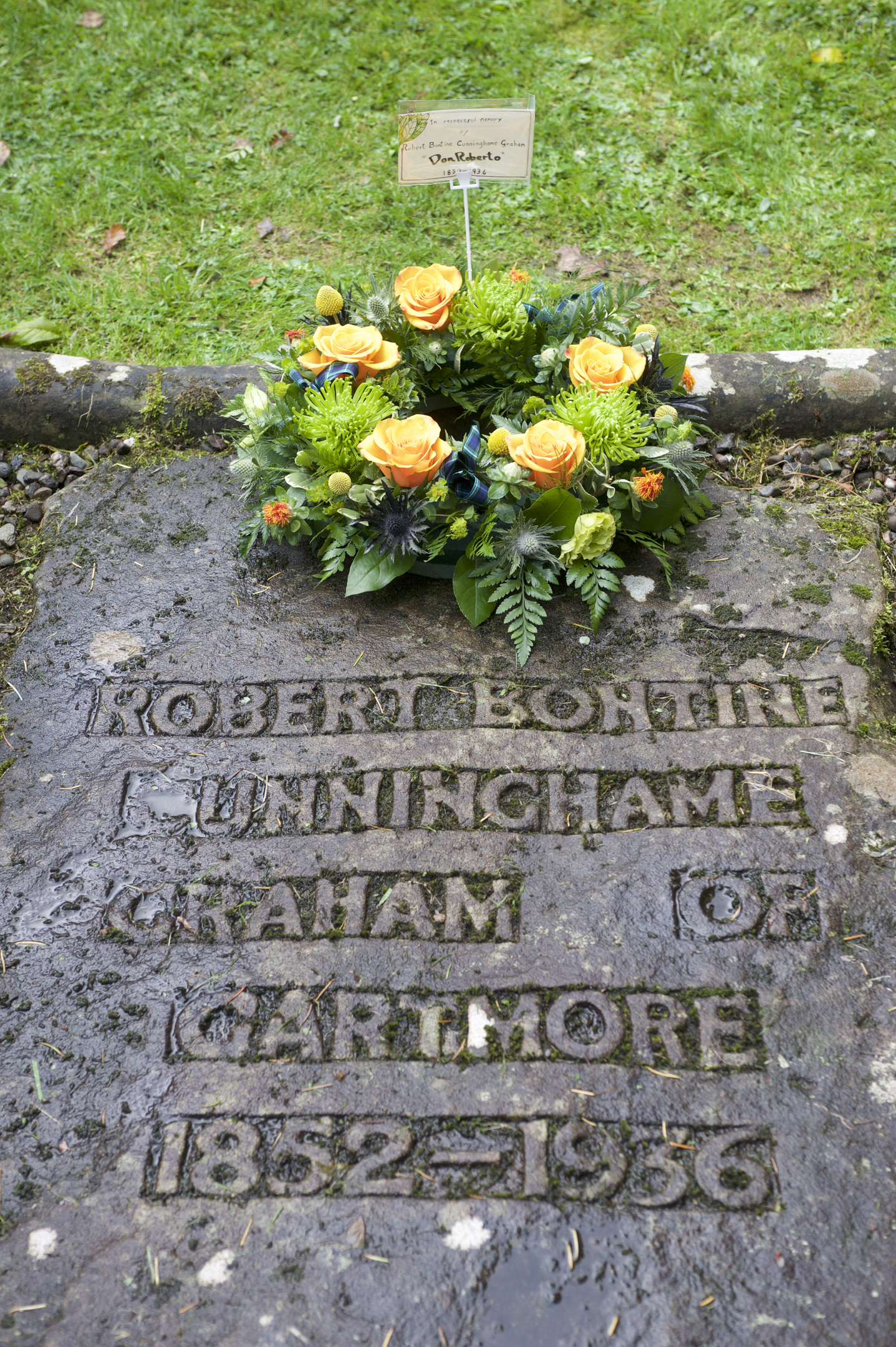 A stone grave slab with engraved name and dates for Robert Cunninghame Graham. It is adorned with a wreath of orange roses and greenery.