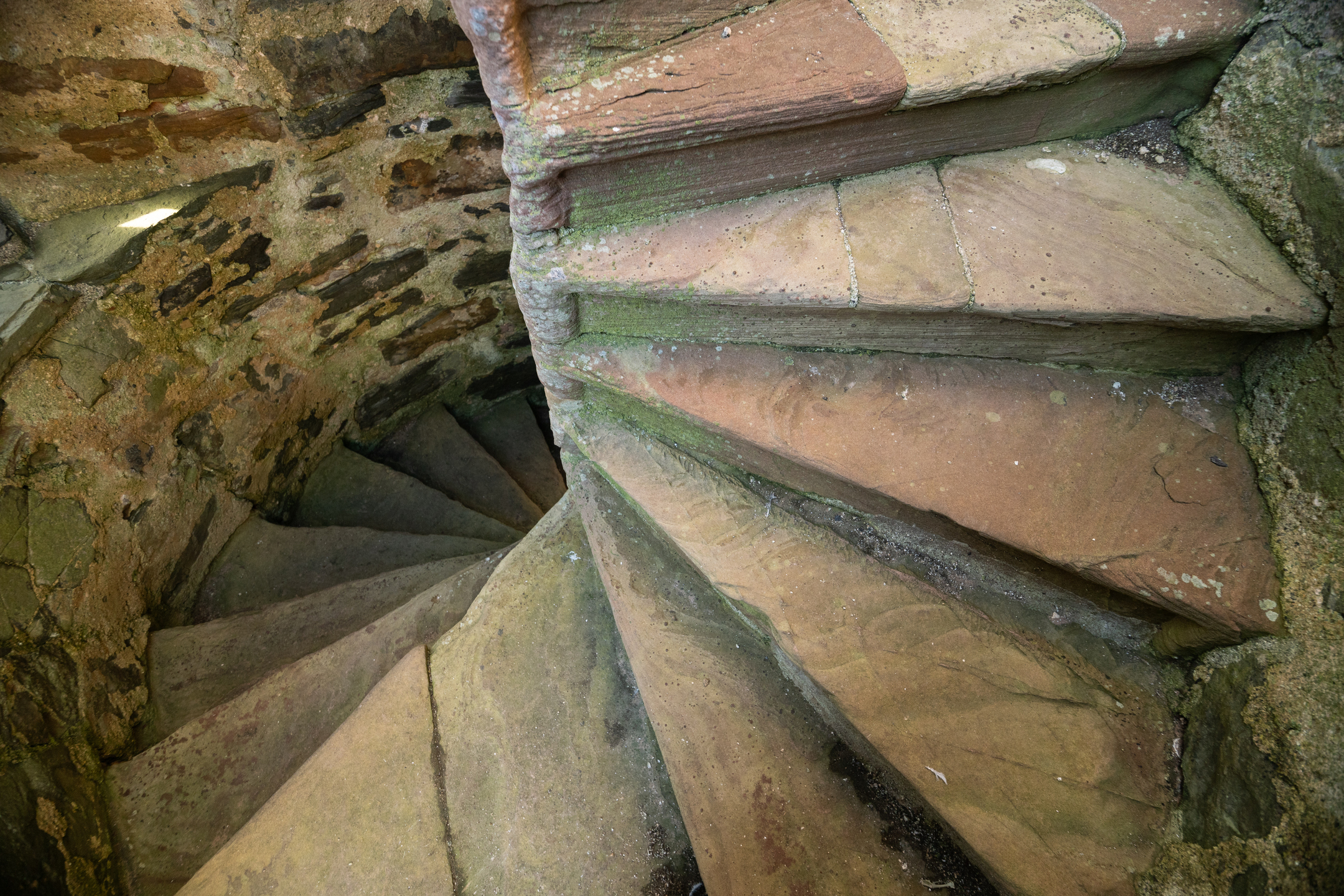 Aerial close-up of a narrow stone spiral staircase, showing deeply worn treads and rough surrounding masonry.