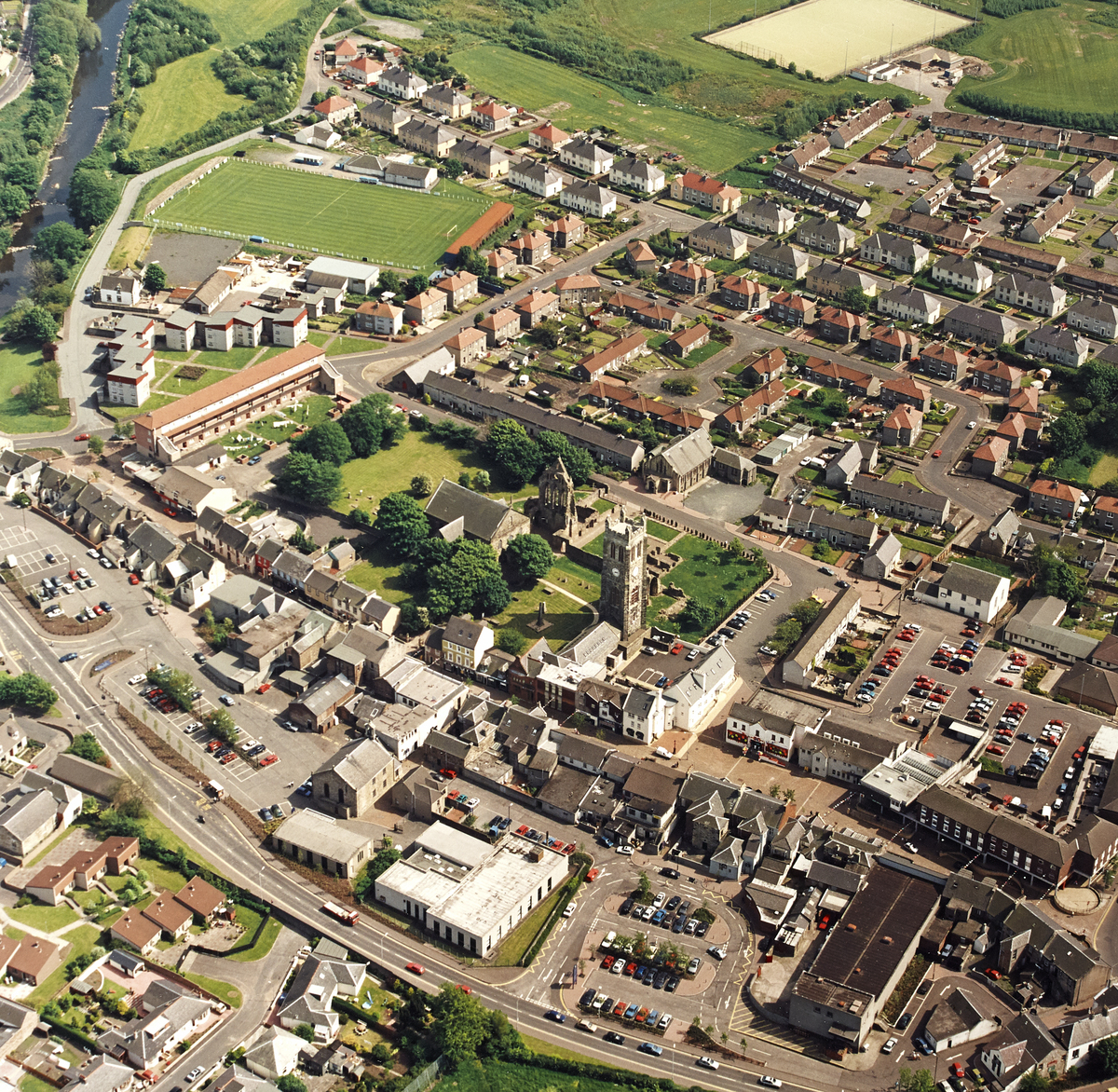 High aerial view of a town showing a large historic church and surrounding ruins at the centre. The church sits within a green open area with trees, bordered by residential streets, car parks, and modern buildings. Rows of houses, a sports field, and a river are visible around the edges of the town.