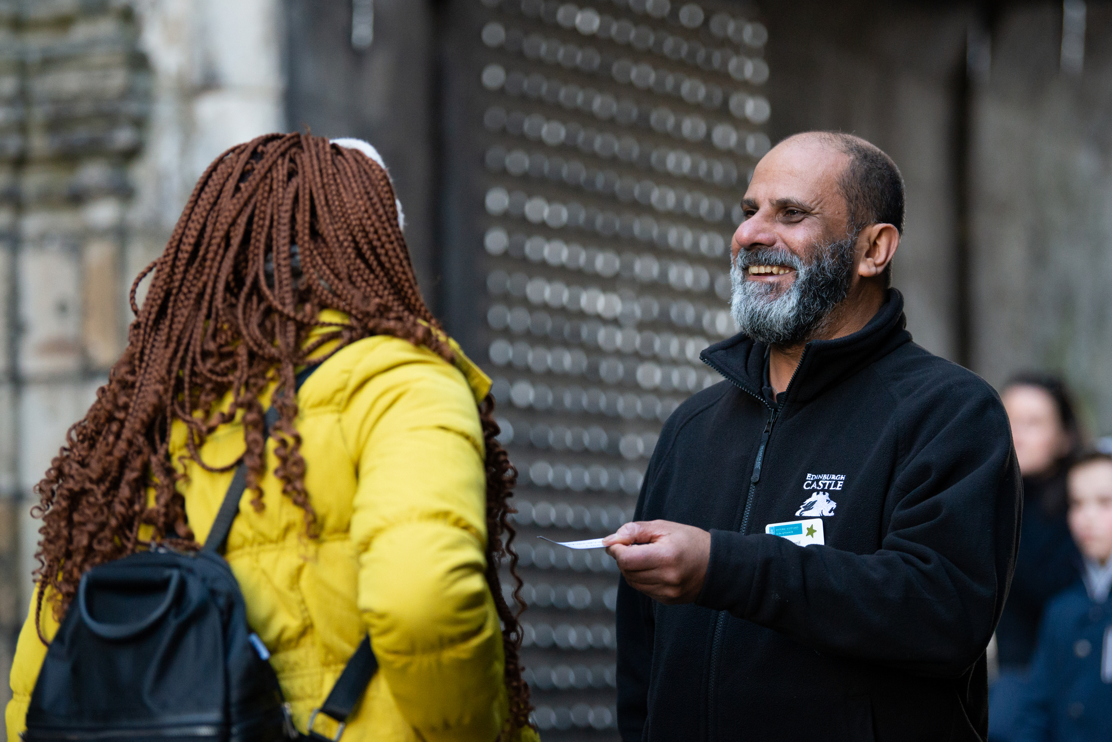 A male admissions assistant wearing an Edinburgh Castle braded jacket handing a ticket to woman in a yellow jacket