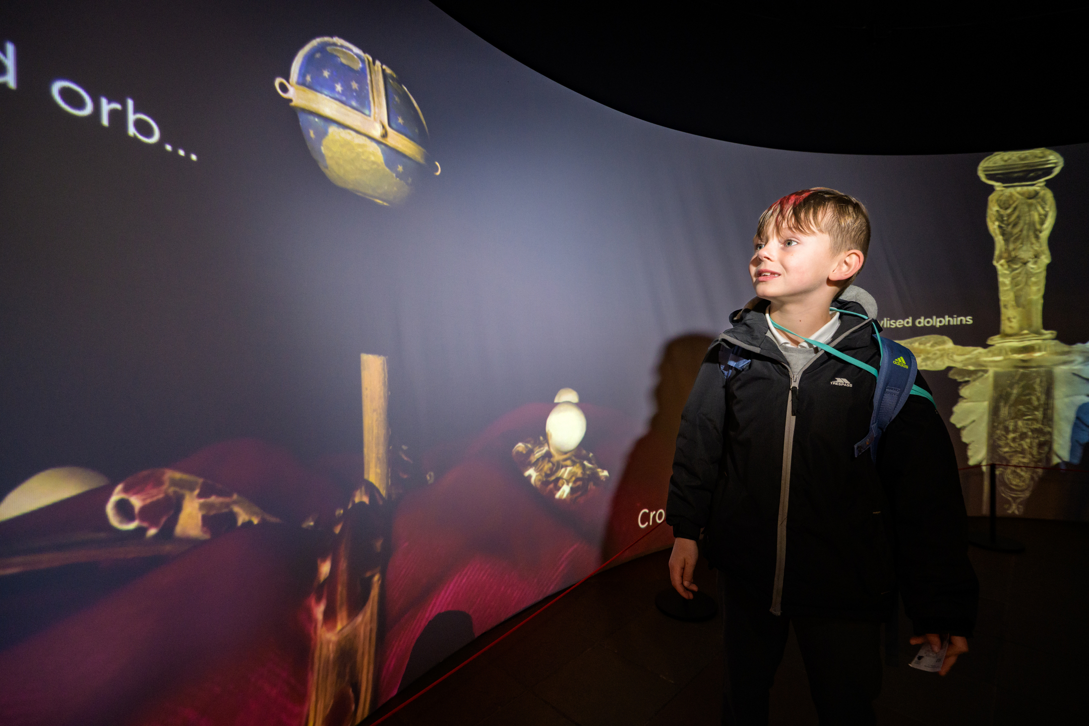 A young visitor stands inside an immersive museum exhibition surrounded by a curved projected display featuring images of historic artefacts, including a ceremonial orb and other treasures. The darkened gallery is illuminated by the large projection, which fills the wall with colourful visuals and interpretive text. The visitor wears a backpack and looks towards the display, engaging with the interactive heritage experience.