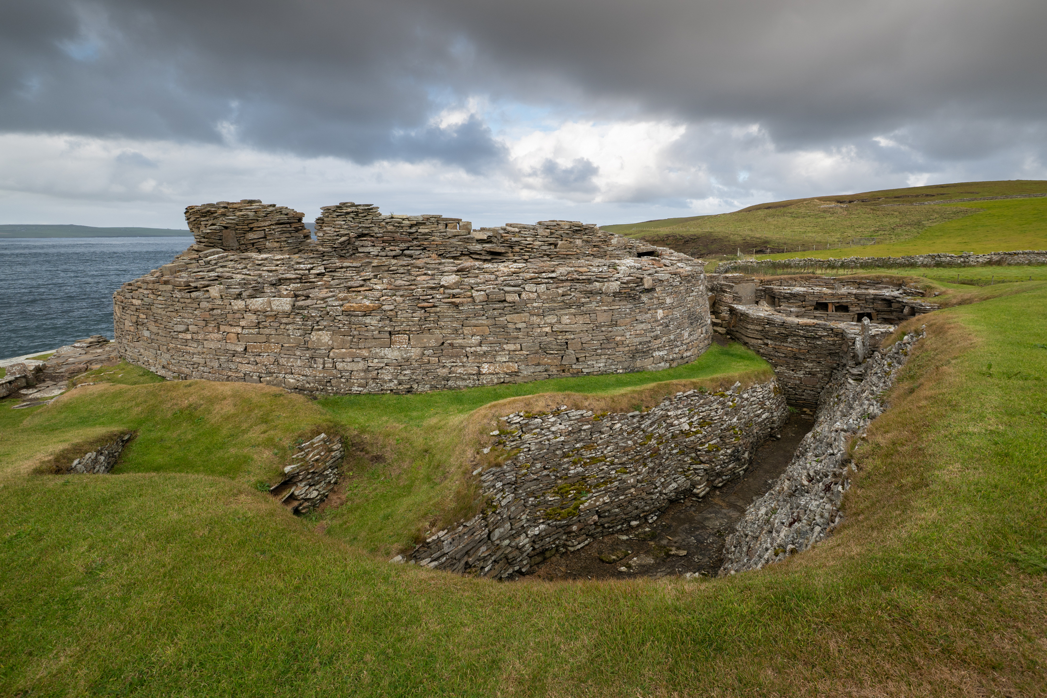 Exterior view of Midhowe Broch’s circular drystone walls under a moody sky, with surrounding grassy banks and the sea behind.