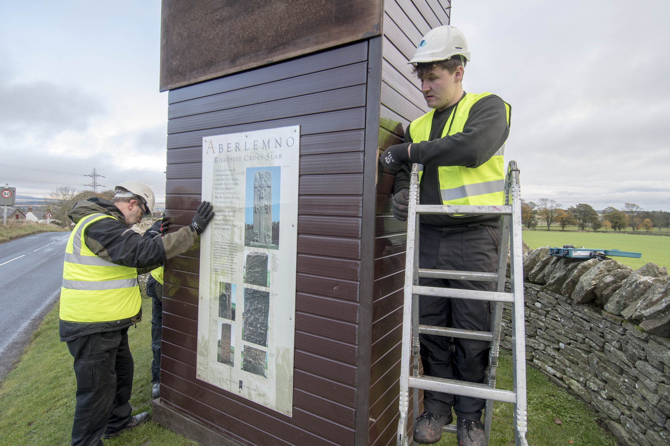 Two workers in hi-vis jackets use a stepladder to place a wooden prot4ective box around a standing stone.