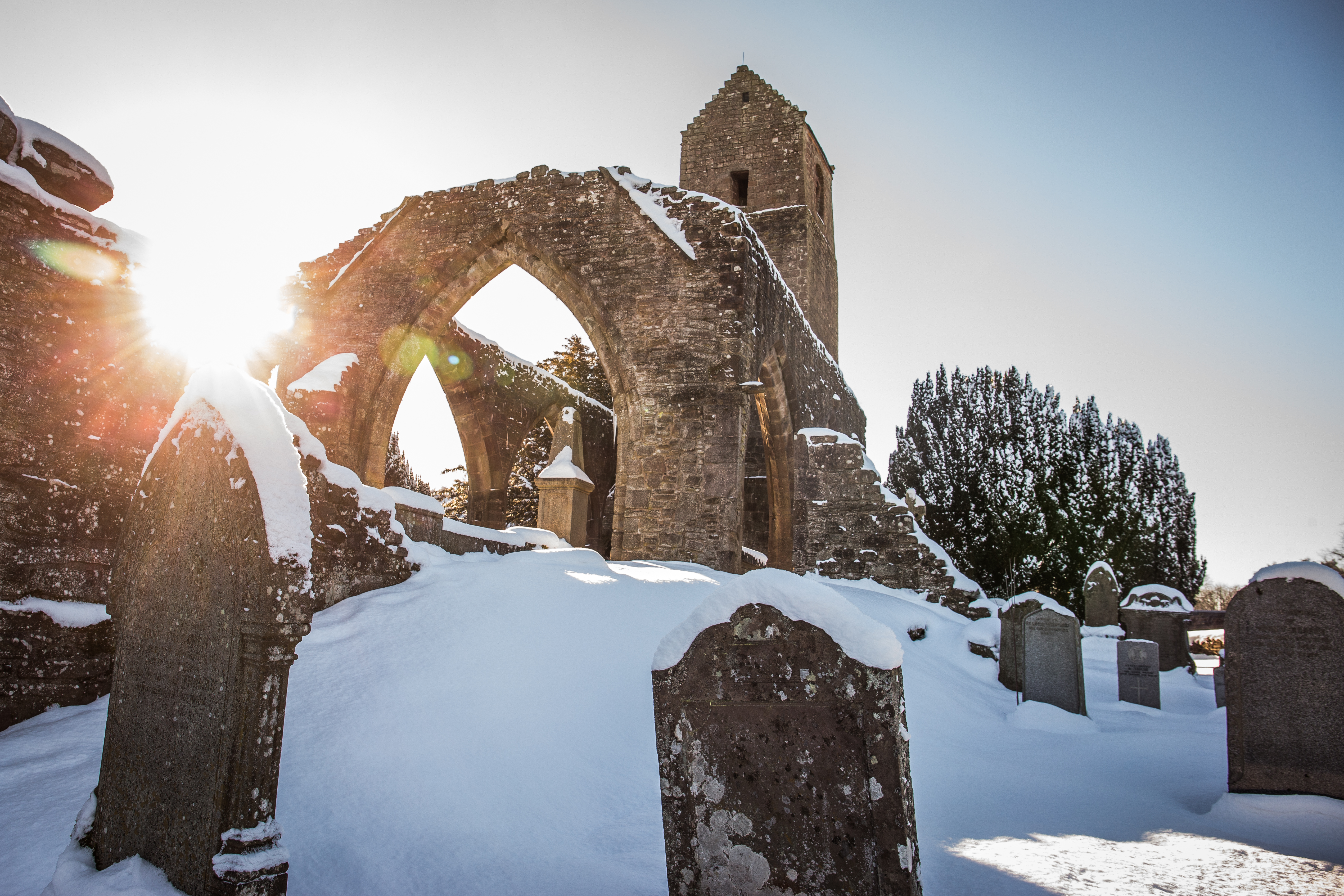 Snow-covered gravestones in front of the arched ruins of Muthill Old Church, with bright sunlight shining through the stone arches.