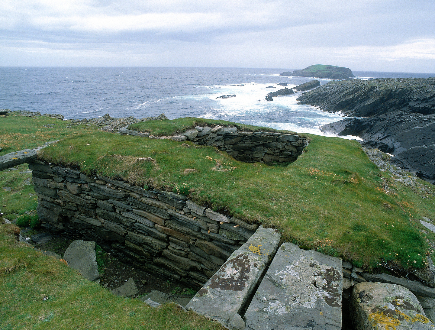 Stone built structure in the ground, with grass on top. There is the sea and cliffs in the background. 