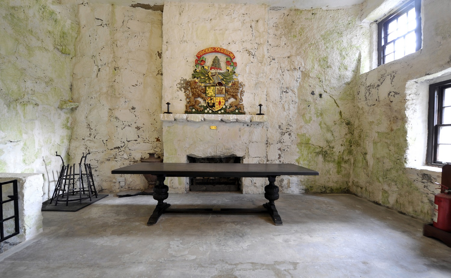 A simple stone-walled room with a large wooden table in the centre, an empty fireplace, and a colourful coat of arms mounted above the mantel.