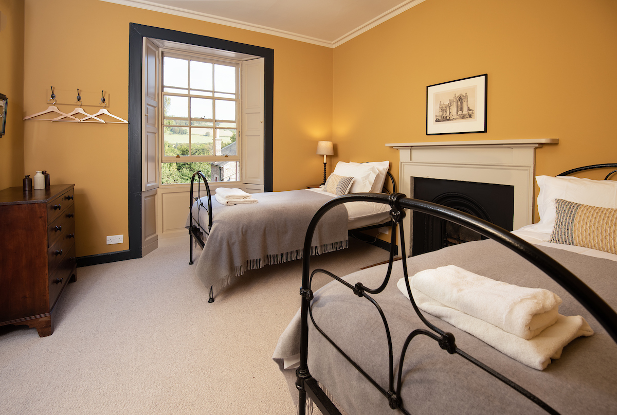 A twin bedroom with mustard walls, a large sash and case window, fireplace, and two black metal beds either side