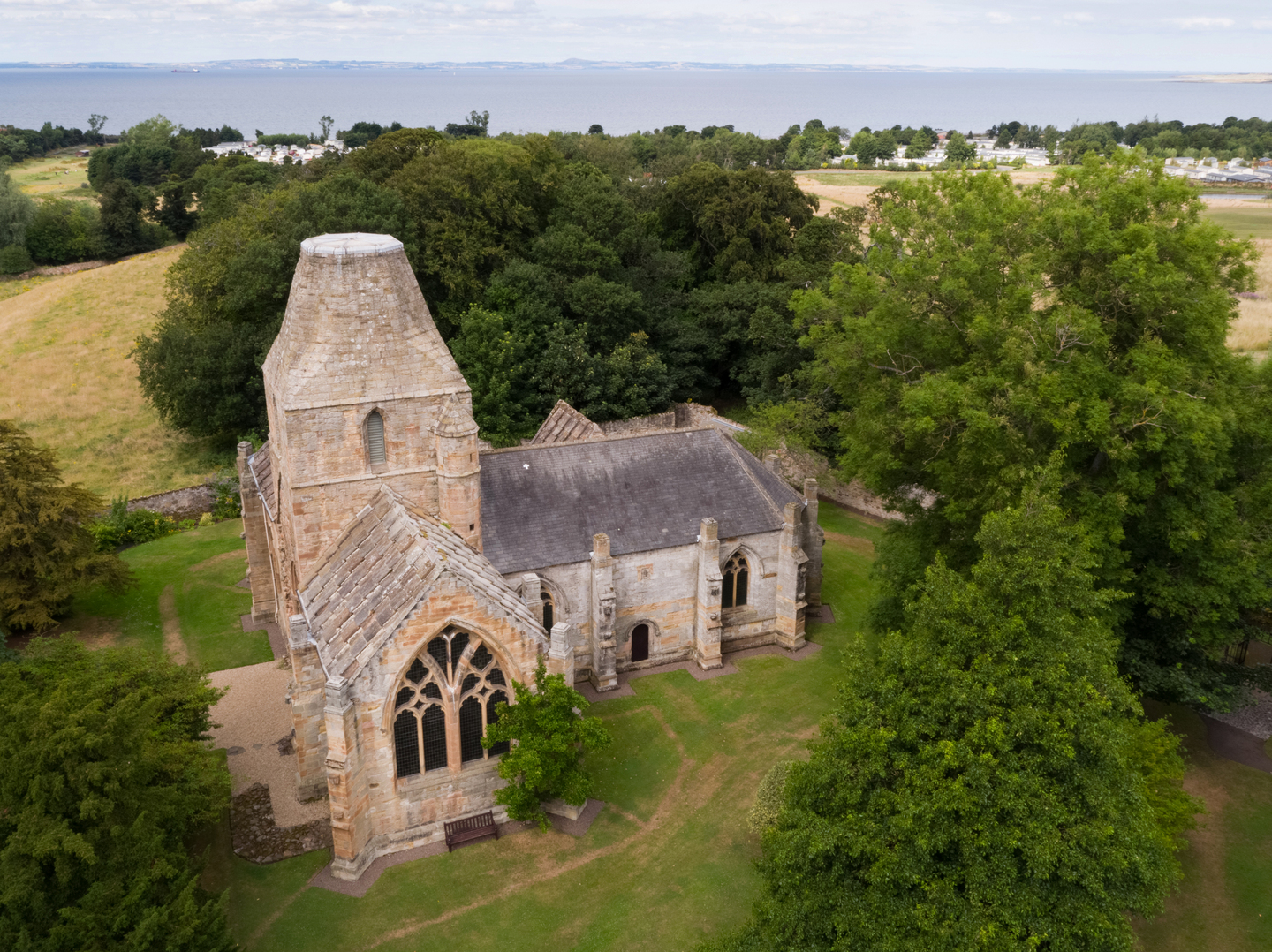 Aerial view of a stone church on a grass landscape, in an L shape. There is a large arched window, with a bench below it. There are trees around the church, and see in the background.