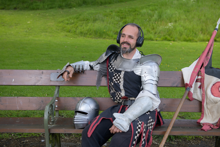A reenactor dressed as a knight sits on a bench and wears headphones. He is smiling and looks relaxed.