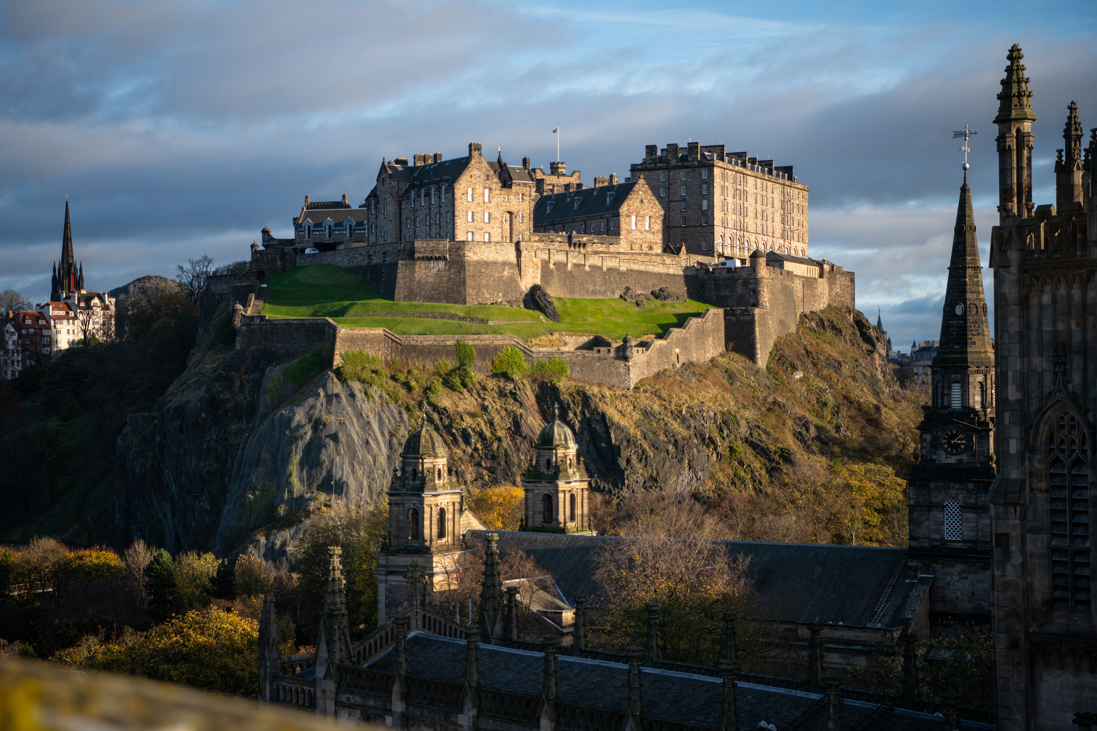 Edinburgh Castle illuminated by sunlight on Castle Rock, with historic rooftops and church spires in the foreground.