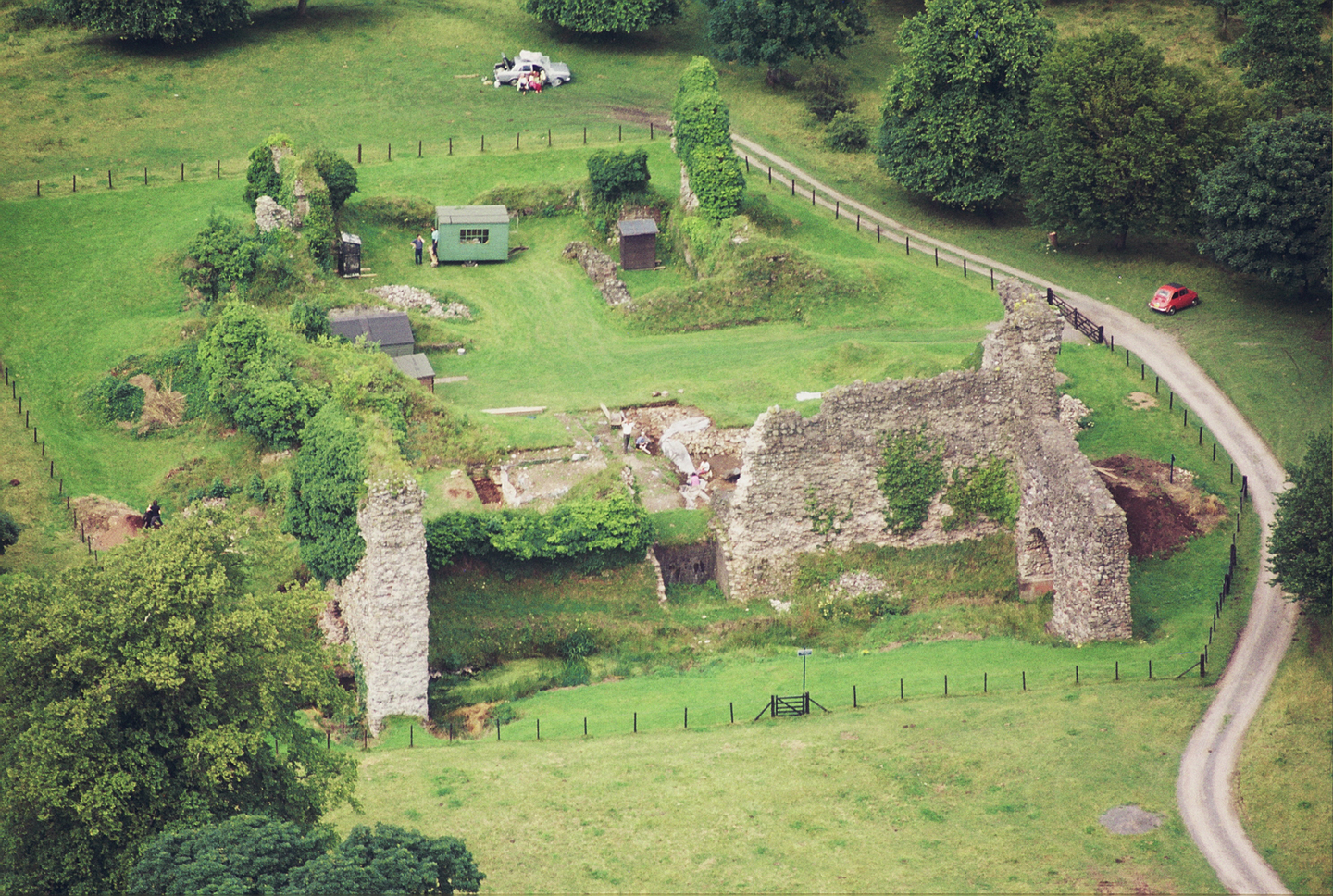 Stone ruins that resemble a castle, there is a wide gate surrounding it, with a road on the right next to it. There are trees and a couple of cars in the background. 