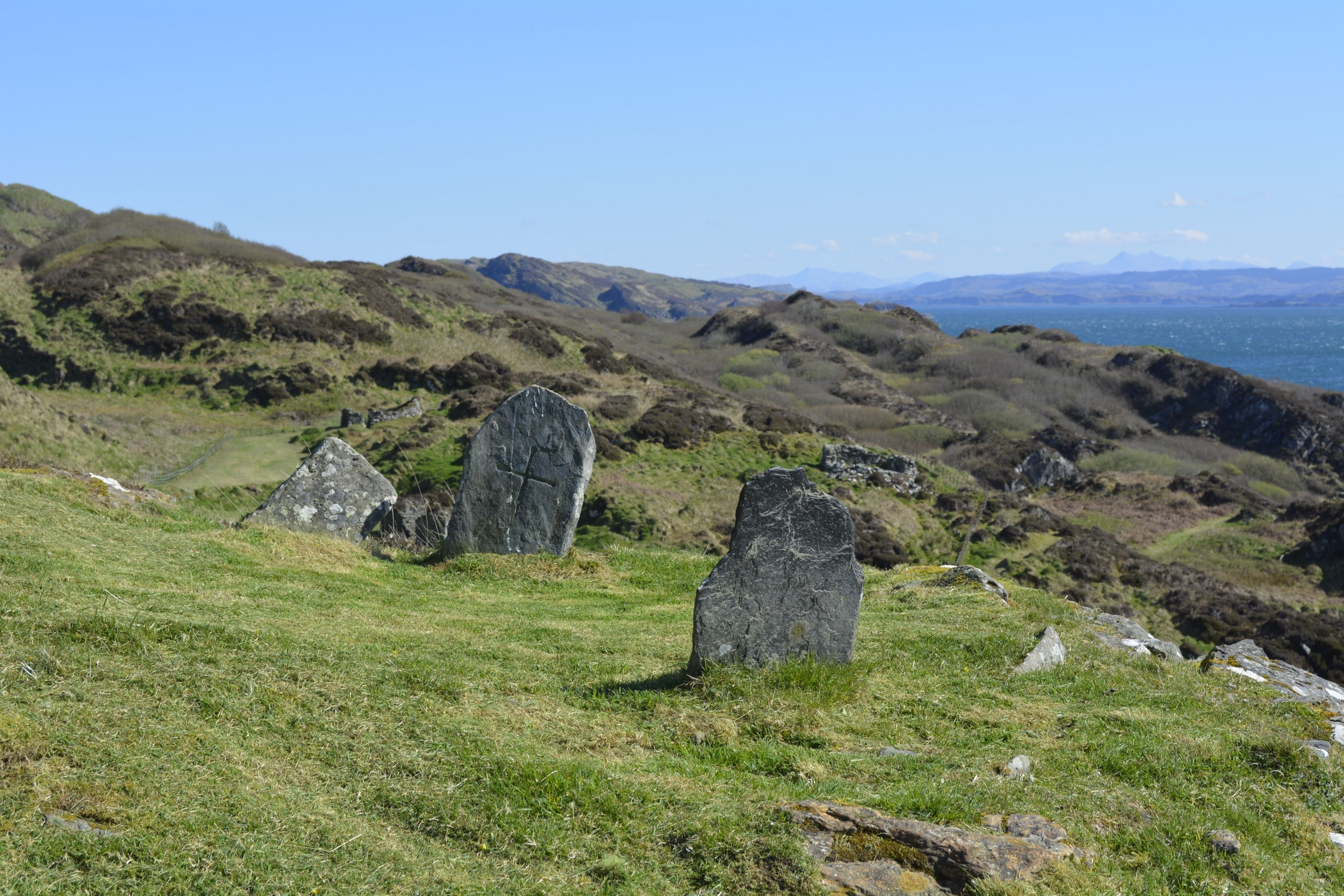 Two weathered stone grave markers standing on grassy ground overlooking rolling hills and the sea under a clear blue sky.