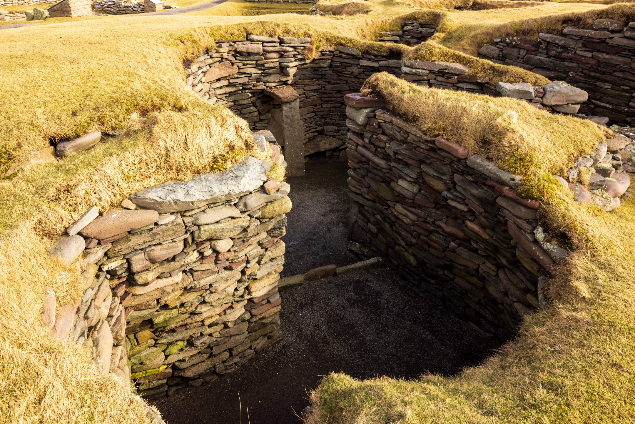 Clearly dug out remains of former building, forming a stone-lined, curved trench in a grassy area