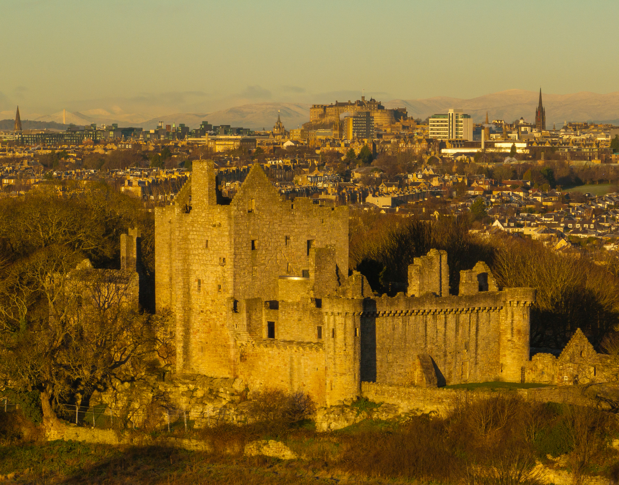 A close elevated view of Craigmillar Castle with sunlit stonework and the city rising behind it.