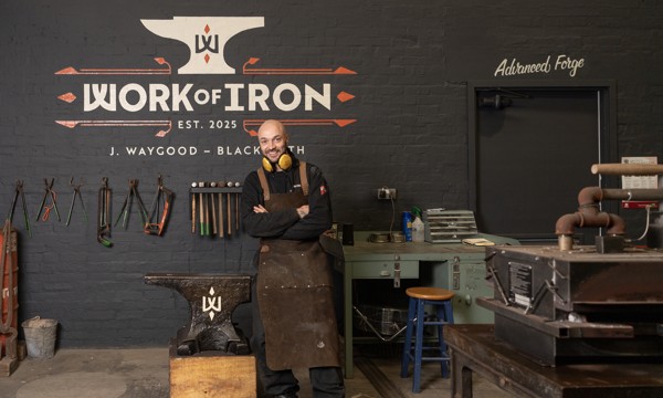 A blacksmith wearing a leather apron and ear protection stands with arms crossed in a forge named 'Work of Iron.' Behind him, blacksmithing tools hang on the wall, and a large anvil with the workshop’s logo is next to him.