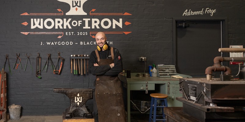 A blacksmith wearing a leather apron and ear protection stands with arms crossed in a forge named 