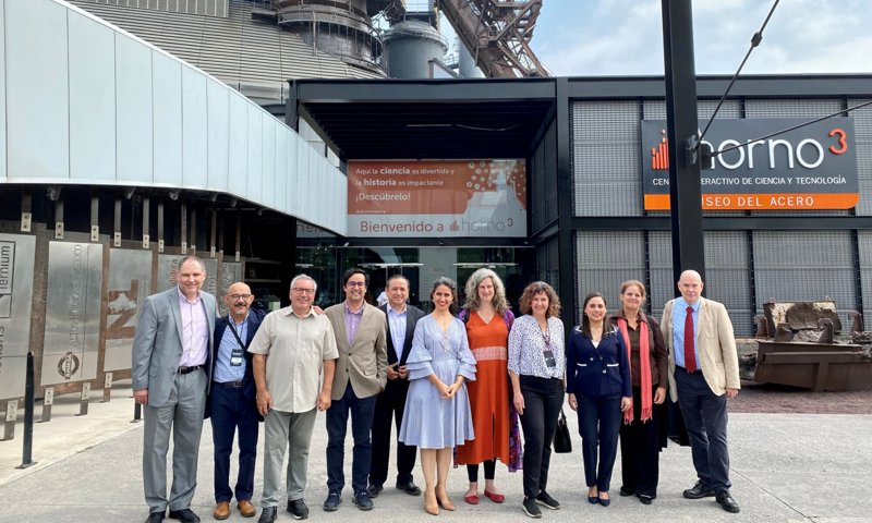 A group of twelve people stand in a line posing for a photograph in front of an industrial museum. The background is a large metal smelter, several stories high, and signage for the museum.