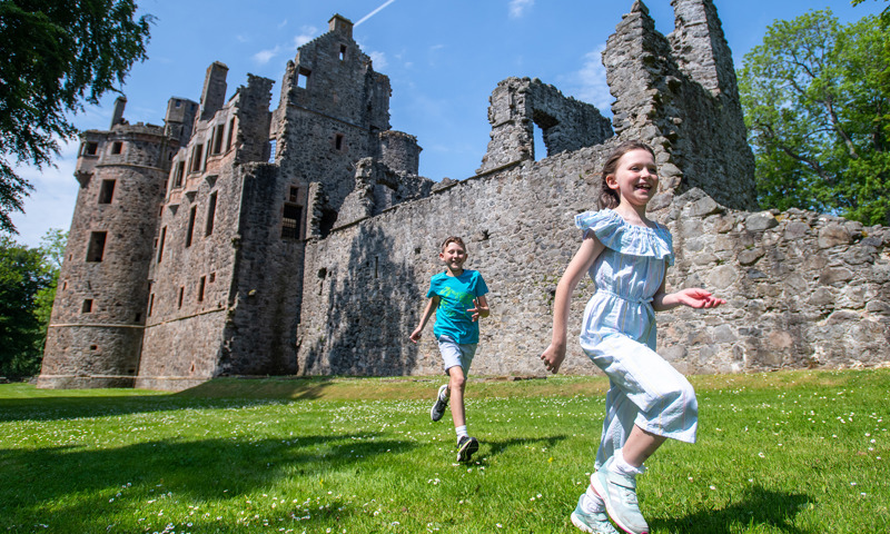 Two children, a girl and a boy, run through across the grassy grounds of an old castle that is in the background. It is a summer day, with blue sky and daisy across the green grass,