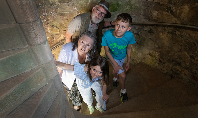 Two children and their grandparents climb a spiral staircase and peer up around the corner.