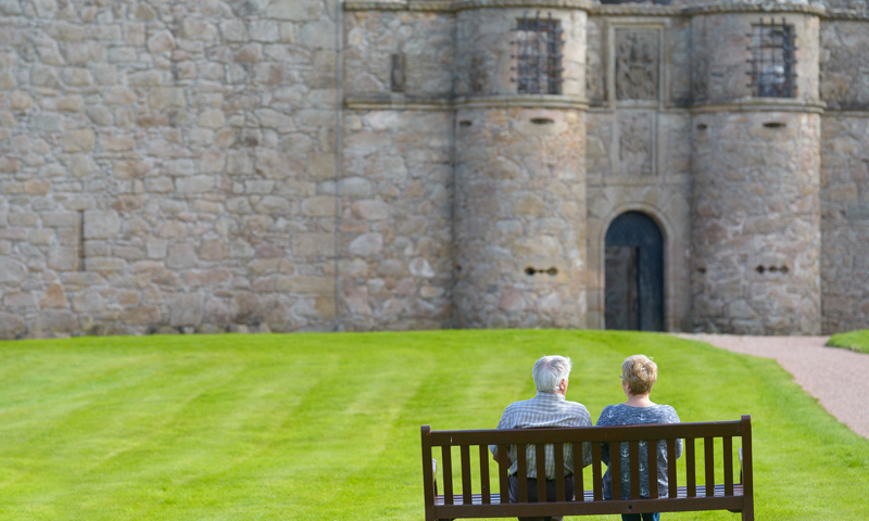 A man and a woman sit on a bench facing towards a castle entrance. The castle is in the background of the image and green freshly cut grass leads from the bench to the entrance, with a gravel path on the right.