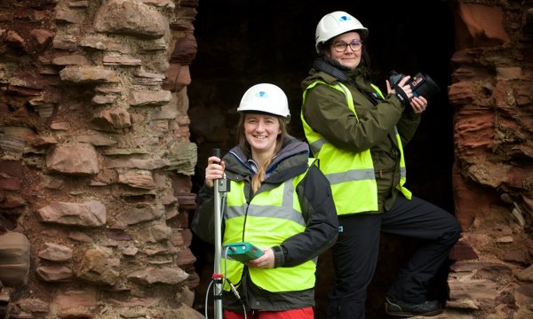 Two people standing in the eroded doorway of an old castle, wearing high vis workwear and holding digital recording equipment.