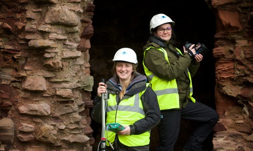 Two people standing in the eroded doorway of an old castle, wearing high vis workwear and holding digital recording equipment.