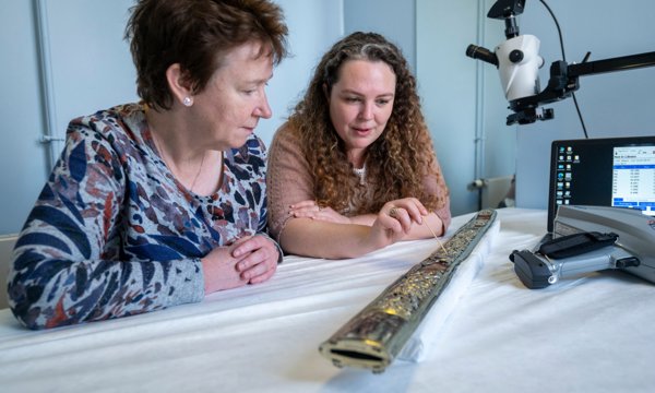 Two people sitting at a table look at a historical scabbard, with scientific recording equipment nearby.
