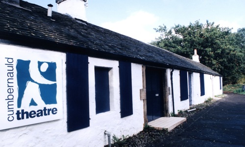 The exterior of a long, one-storey whitewashed cottage, with a black door and black wooden shutters on the window. A modern sign near the entrance tells us it is Cumbernauld Theatre.
