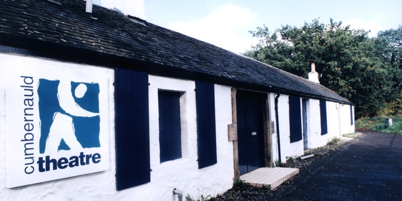 The exterior of a long, one-storey whitewashed cottage, with a black door and black wooden shutters on the window. A modern sign near the entrance tells us it is Cumbernauld Theatre.