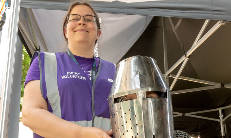 A person in a purple volunteer jacket with a child in a helmet and chainmail 