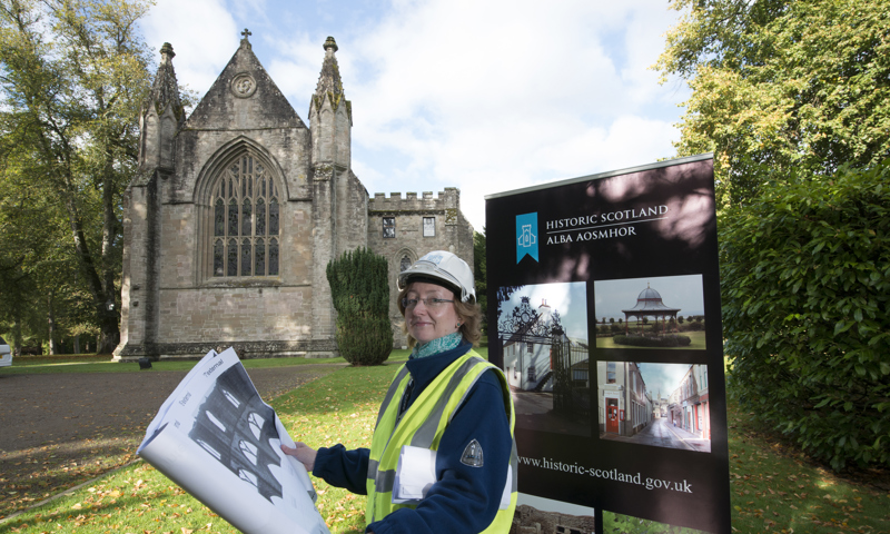 Female architect holding plans, standing in front of a stone cathedral with an interpretation panel behind her.