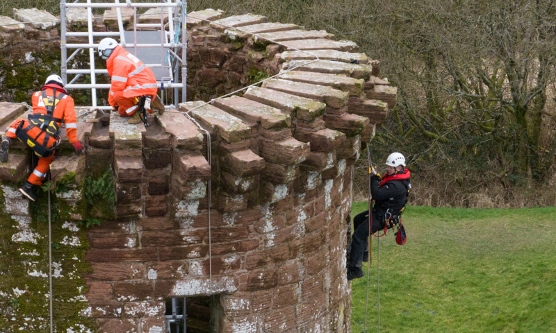 Three people using scaffolding and ropes to do inspection work at the top of a tower of Caerlaverock Castle.