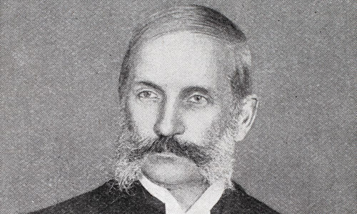Black and white portrait photo of a young looking John Thomson from 1898. Thomson has a mutton-chop style beard, with his long dark moustache joining up with even longer white sideburns. He is wearing a dark suit, with a white shirt and tie.